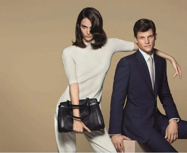 A woman in a white ribbed dress holding a black handbag poses with a man in a navy suit.