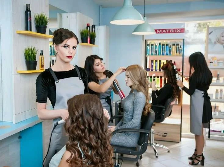 Stylists in grey aprons work on clients' hair in a bright salon featuring blue and wood accents.
