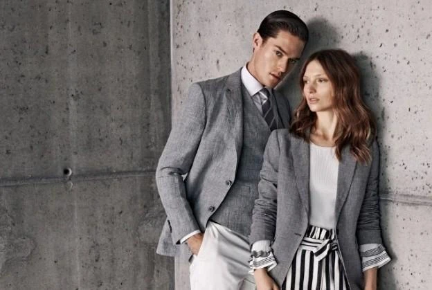 A man and woman leaning against a concrete wall wearing matching light grey textured blazers.