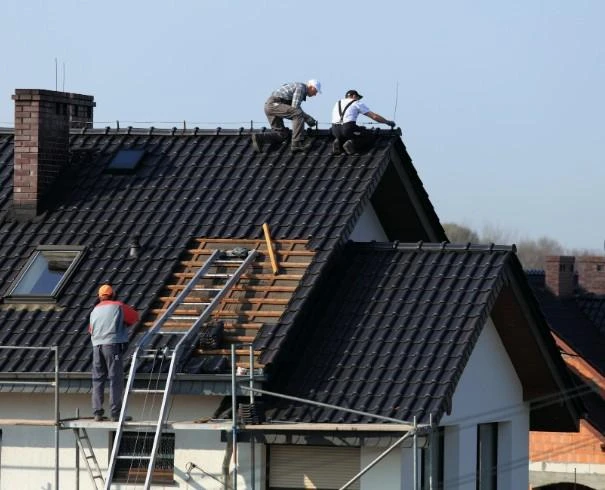 Roofers working on a dark tiled roof with a ladder and scaffolding visible on a white house.