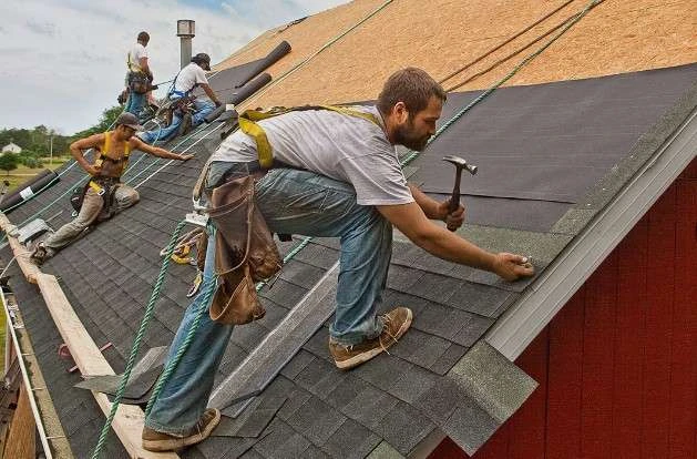 A team of construction workers wearing safety harnesses installs shingles on a sloped roof.