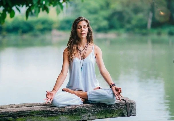 A woman in white clothing sits in a lotus meditation pose on a wooden beam over a calm lake.